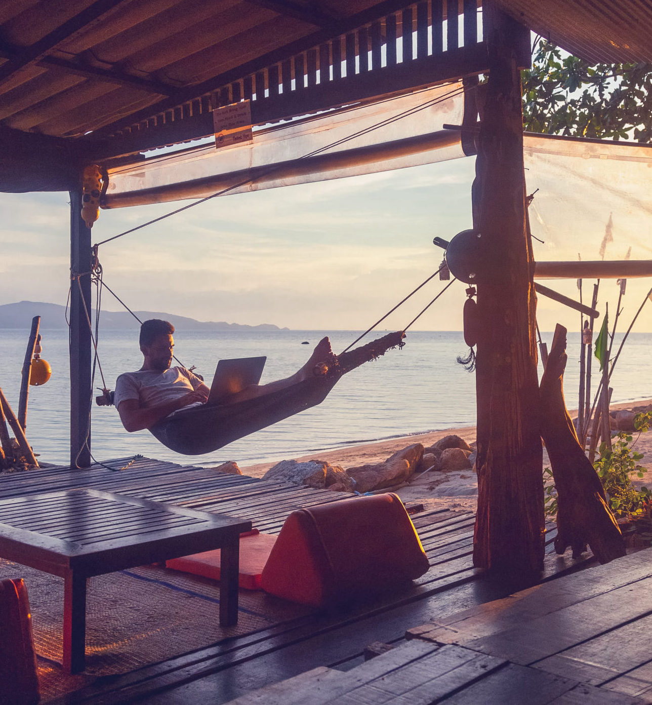 a man with a beard in a hammock with a laptop on the background of the sea and sunset, distant work, freelancer, blogger, vacation and travel